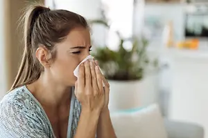 A woman is sitting on a couch and blowing her nose with a tissue while looking down.
