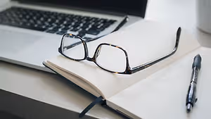 A desk with a laptop, an open notebook, a pen, and a pair of glasses.