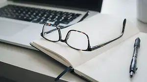 A desk with a laptop, an open notebook, a pen, and a pair of glasses.