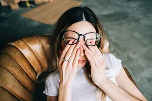 A woman wearing glasses is sitting on a brown chair while covering her eyes with her hands