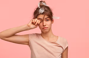 A woman with brown hair and a pink shirt is posing for a photo with feathers in her hair while closing her eyes with her hand on her forehead.