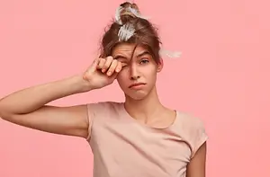 A woman with brown hair and a pink shirt is posing for a photo with feathers in her hair while closing her eyes with her hand on her forehead.