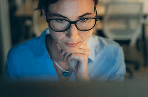 A woman with glasses and a necklace is looking at a laptop screen with a serious expression