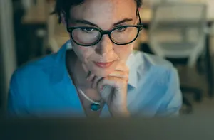 A woman with glasses and a necklace is looking at a laptop screen with a serious expression