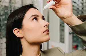 A woman with brown hair is looking upward and seems to be applying eye drops. She is wearing a brown coat and has black nail polish. She is holding a white bottle in her right hand. Behind her is a glass window with a white frame and a white door.