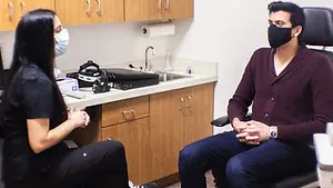 A man wearing a face mask is sitting in a chair and talking to a woman who is also wearing a face mask and sitting on a chair in front of a sink and cabinets.