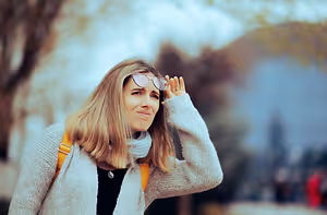 A woman in a gray sweater is standing in an outdoor area, smiling and looking at something while holding her glasses with her left hand and touching her hair with her right hand. The background is blurry, with trees and a building.