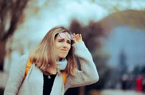A woman in a gray sweater is standing in an outdoor area, smiling and looking at something while holding her glasses with her left hand and touching her hair with her right hand. The background is blurry, with trees and a building.