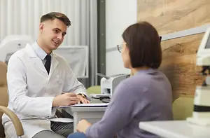 A man in a white coat talks to a woman who is sitting across from him in an office.