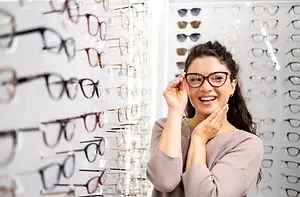 A smiling woman wearing glasses is standing in front of a wall full of eyeglasses, posing for a picture.