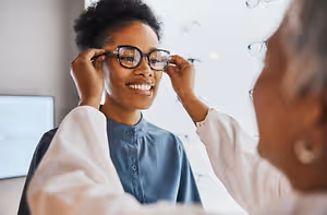 A smiling woman trying on glasses in an optometrist's office with a doctor adjusting her glasses.