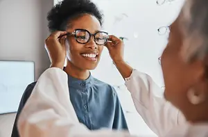 A smiling woman trying on glasses in an optometrist's office with a doctor adjusting her glasses.