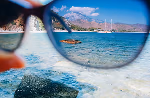 A pair of glasses showing a view of a beach with rocks, water, and mountains in the distance.