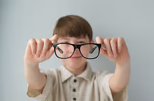 A young boy is holding up a pair of black-framed glasses with a slightly blurry view of the reflection of his face.