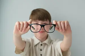 A young boy is holding up a pair of black-framed glasses with a slightly blurry view of the reflection of his face.