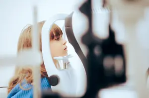 A young girl with blonde hair is having an eye test with a blurry background behind her.
