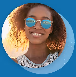 A smiling woman with curly hair and sunglasses standing outside in the sun with a blue background