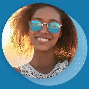 A smiling woman with curly hair and sunglasses standing outside in the sun with a blue background