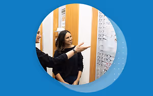 Two women are smiling and looking at glasses in a store. One of them is pointing at the glasses and the other one is smiling and looking at her.