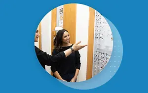 Two women are smiling and looking at glasses in a store. One of them is pointing at the glasses and the other one is smiling and looking at her.