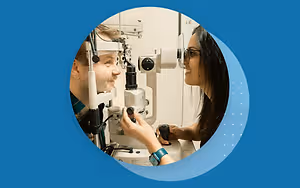 An optometrist is examining a patient's eye using an ophthalmoscope. The patient is wearing eyeglasses and is looking into the machine, while the doctor is operating it.