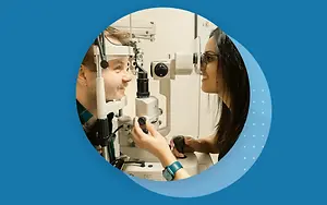 An optometrist is examining a patient's eye using an ophthalmoscope. The patient is wearing eyeglasses and is looking into the machine, while the doctor is operating it.
