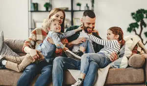 A family is sitting on a couch and smiling for a photo. The father is holding the boy and the boy is holding the girl. The mother is holding the boy. The girl is sitting on the father's lap. The boy is sitting on the couch. The girl is wearing a striped shirt. The boy is wearing a sweater. The parents are wearing sweaters. The couch is covered with a blanket.