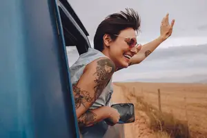 A woman with tattoos is waving her hand from the window of a blue car while smiling and looking forward on a road in the countryside