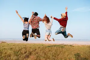 Four individuals, two women and two men, jumping together on a grassy field with blue sky above and a serene beach in the background.