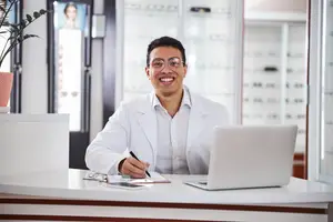 A smiling man in a lab coat sits at a desk with a laptop and a pen, surrounded by a potted plant and shelves with items.