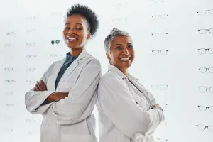 Two women wearing white coats standing in front of a white wall with many pairs of eyeglasses displayed.