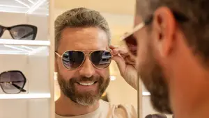 An adult male is adjusting his sunglasses in front of a mirror, smiling and looking at the reflection of his face.
