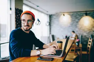 A man in a blue long sleeve shirt and an orange beanie sits at a table in a coffee shop and types on a laptop