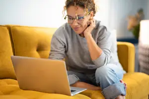 A woman is sitting on a couch and is using a laptop while wearing glasses and a gray long-sleeve shirt.