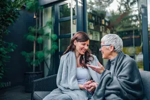 Two women sitting on a patio, one wearing glasses, smiling and talking to each other