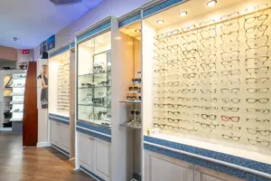 An interior view of an eyeglasses shop featuring a display of glasses on a wall and a cabinet with shelves, a wooden floor, and a woman's picture on the wall.