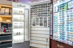 An empty shop with shelves filled with sunglasses and glasses displayed on white shelves with a wall that has many sunglasses on it, a poster of a logo, a wall with a power outlet, a red bottle, a wooden shelf, and a logo on the bottom right.