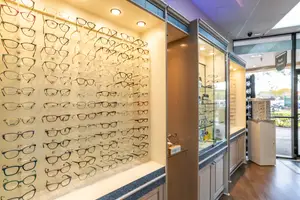 Inside an optical shop, many frames hang on a wall in an organized manner, and a glass display case is on the right side. A box is placed on the counter, and a glass door is on the right side. A surveillance camera is mounted on the ceiling. A car is parked outside the shop.