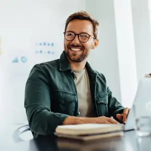 A smiling man sitting at a desk in front of a laptop with a book on the table