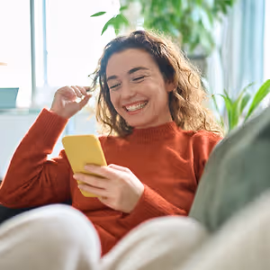 A woman smiling while looking at her phone with one hand holding her glasses