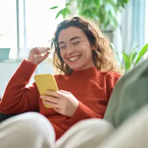 A woman smiling while looking at her phone with one hand holding her glasses