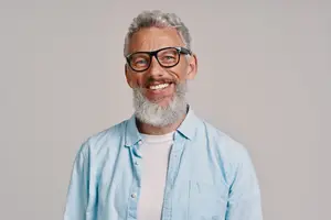 A smiling man with gray hair and a beard wearing a blue shirt and black glasses