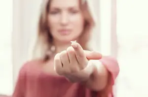 A woman wearing a pink shirt is pointing to a contact lens she is holding in her hand.