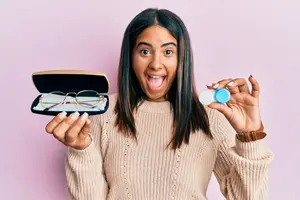 A woman wearing a sweater is holding a pair of glasses and a contact lens bottle in her hands.