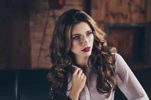 A woman with long brown wavy hair wearing a white shirt and glasses is sitting on a black couch.