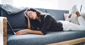 A woman is lying on a couch while using a laptop with her hand on her head. She is wearing glasses and white socks. Behind her is a gray blanket on the couch. The couch is placed on a white wall, and a white pillow is on the right side of the couch. A wooden table is placed on the left side of the couch.