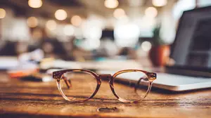 A pair of glasses on a desk with a laptop and other objects in the background