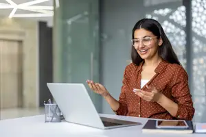A woman sitting at a desk with a laptop and smiling, possibly in an office setting.