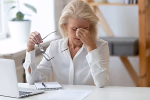 An older woman with glasses sits at a desk with a laptop and papers, looking distressed