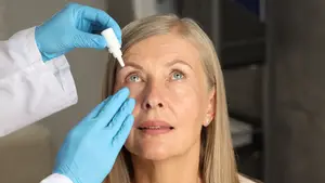 An elderly woman with blonde hair is having her eyes examined by a doctor wearing a white coat and blue gloves.